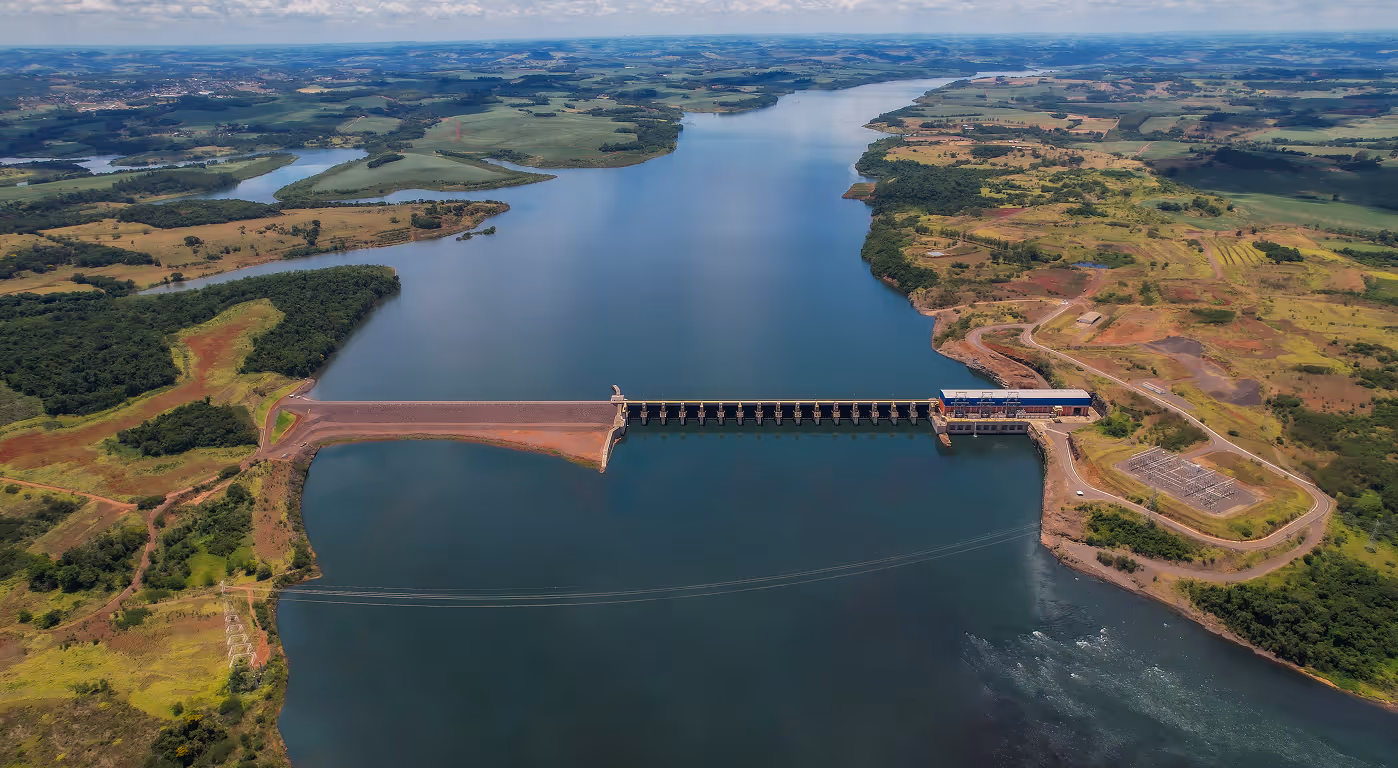 Aerial view of a large dam spanning a wide river surrounded by green and brown fields under a partly cloudy sky.