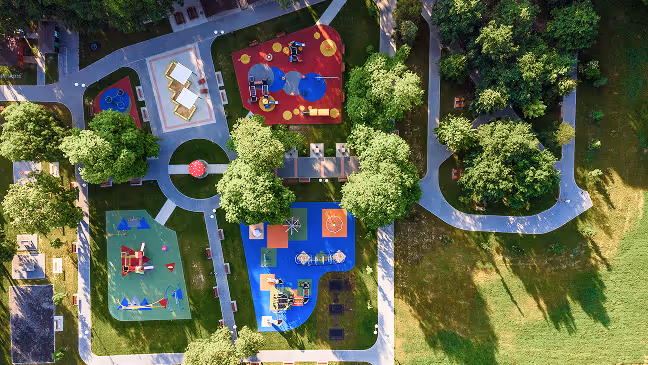 Aerial view of a colorful playground with slides and climbing structures surrounded by trees and intersecting walking paths in a green park.