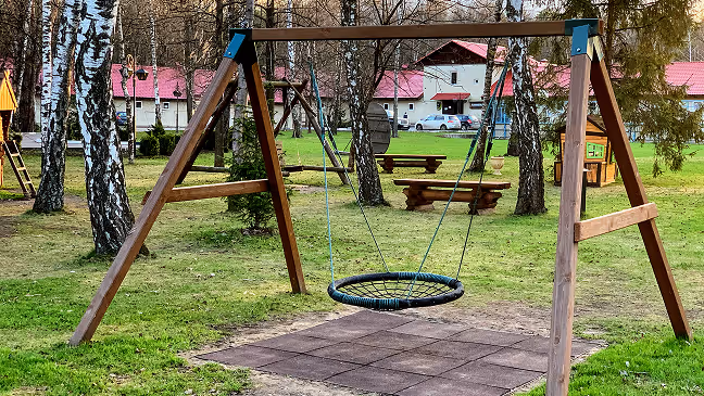 A round rope swing hanging from a wooden frame in a grassy playground with birch trees and picnic tables in the background.