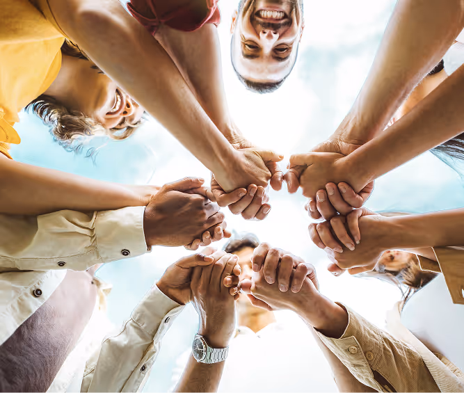 Group of diverse people standing in a circle holding hands together with a bright sky background.