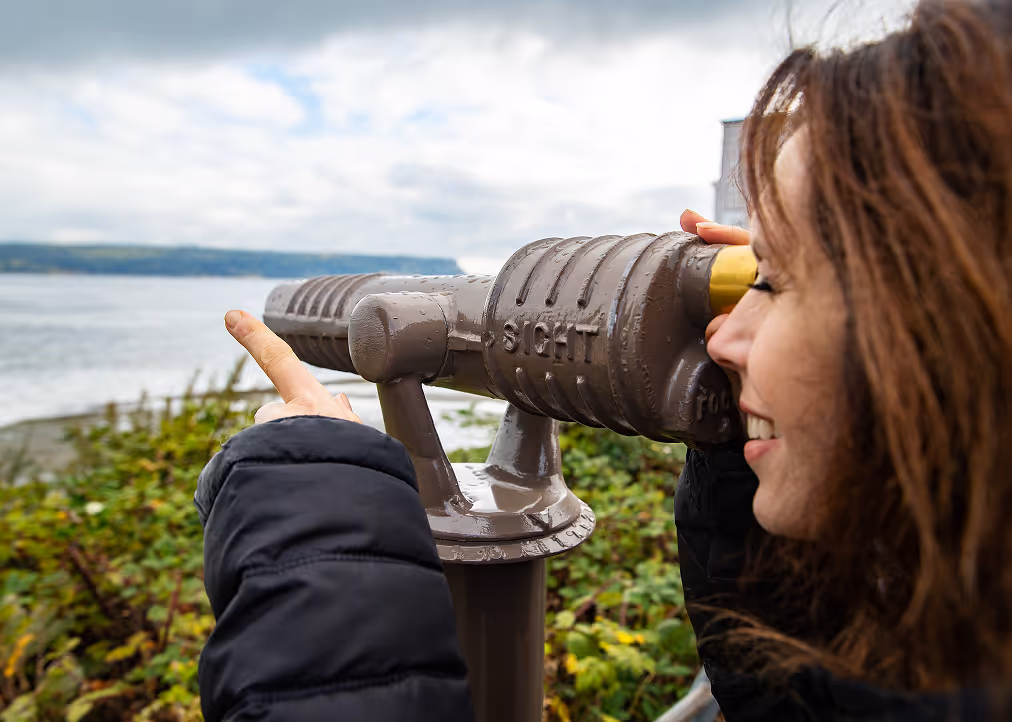 Smiling woman looking through a mounted sightseeing telescope near a body of water with green foliage in the background.