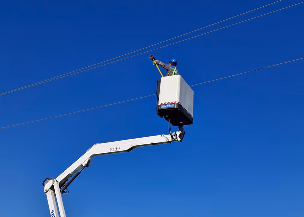 Worker wearing a blue helmet and safety harness elevated in a bucket lift working on overhead power lines against a clear blue sky.