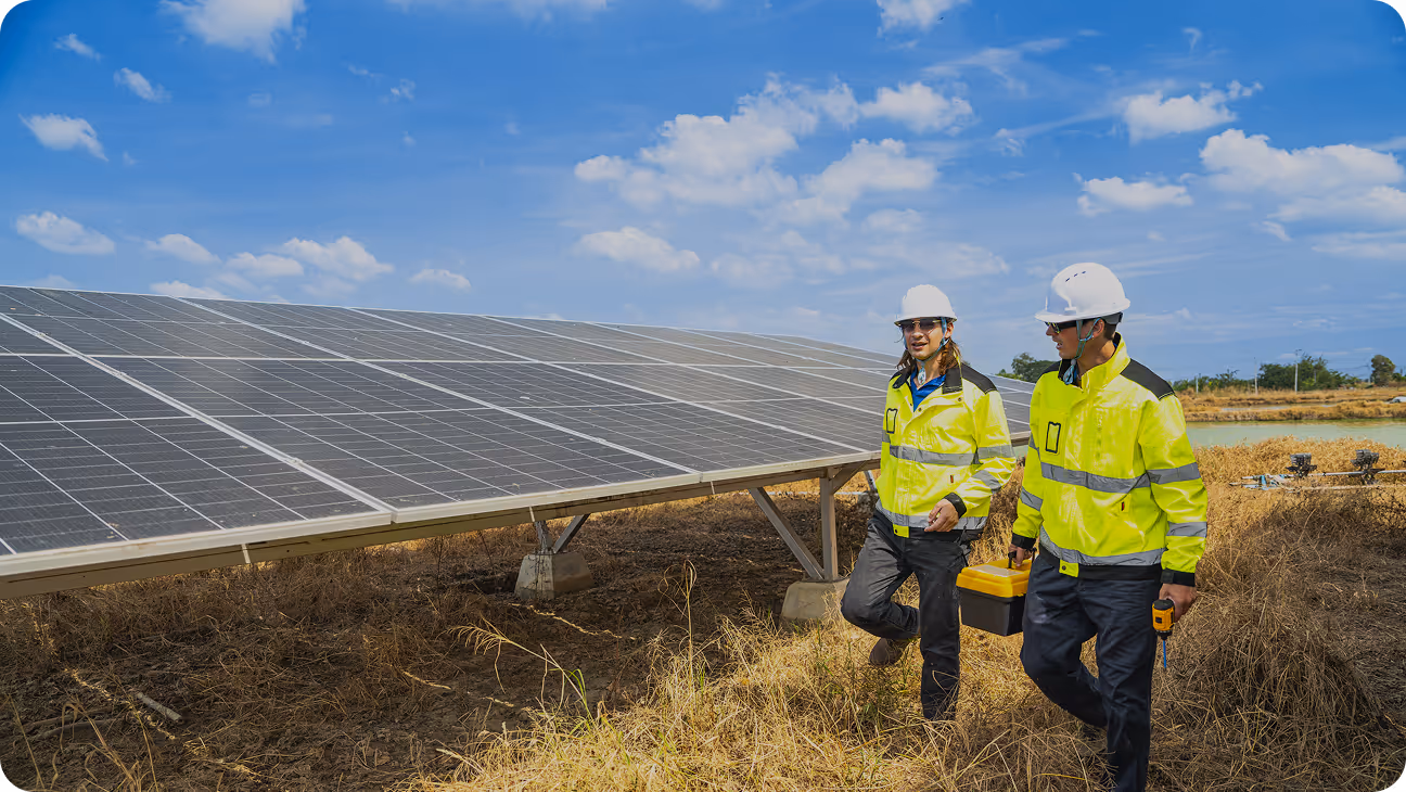 Two workers in yellow safety jackets and white helmets inspecting solar panels outdoors under a blue sky.