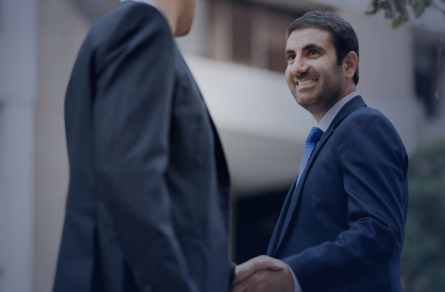 Two businessmen shaking hands, one smiling and wearing a blue suit and tie.