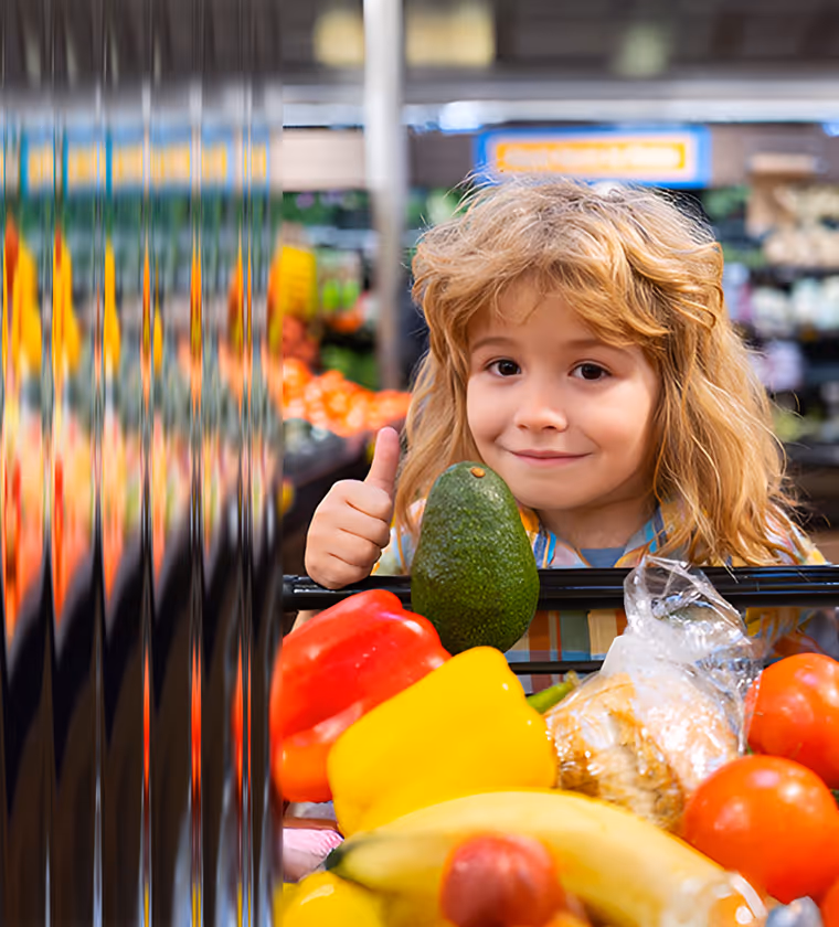 Smiling child holding an avocado and giving a thumbs up behind a shopping cart filled with colorful fresh vegetables and fruits in a grocery store.