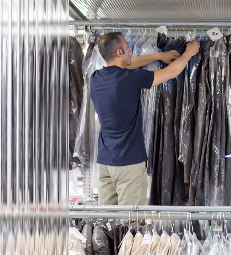 Man arranging dry-cleaned clothes covered in plastic hanging on a rack in a laundry facility.