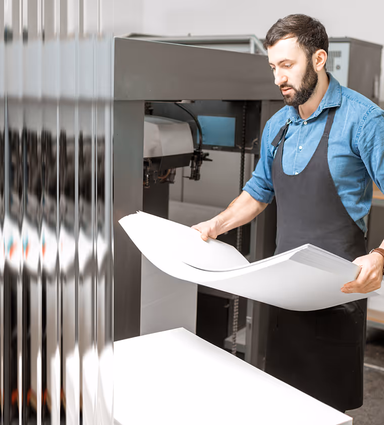 Man in a blue shirt and black apron holding large sheets of paper next to a printing machine.