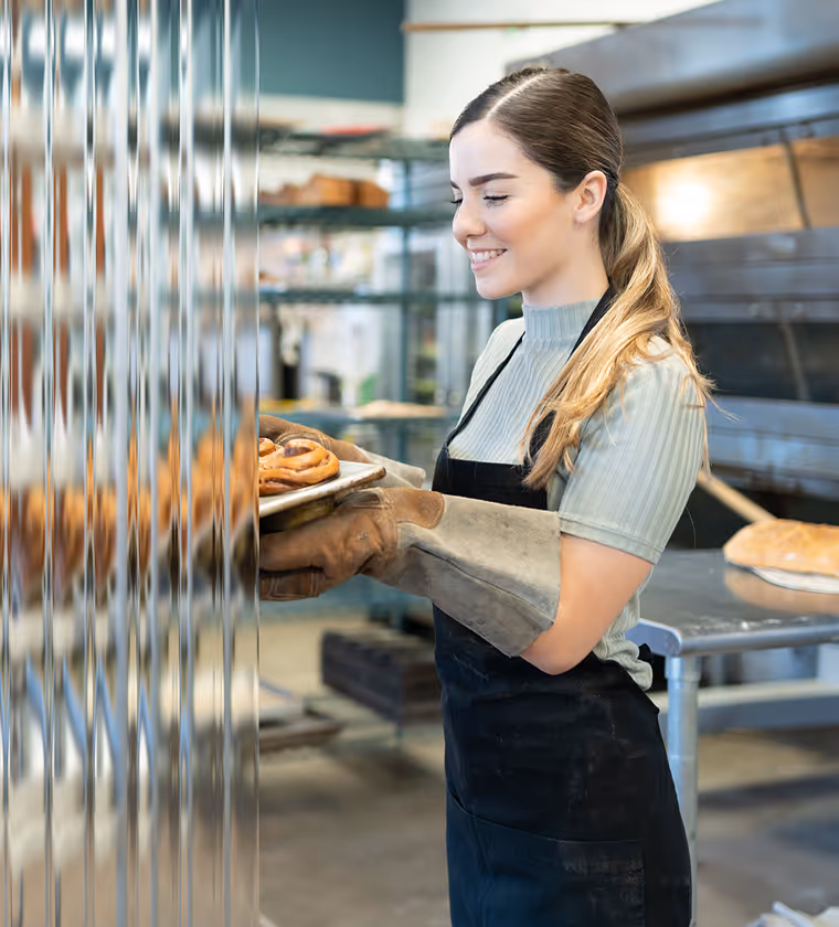 Smiling woman wearing oven mitts and apron placing a tray of freshly baked pretzels into an oven in a bakery.