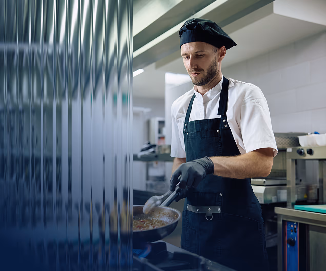 Chef wearing a black apron and hat cooking with tongs in a frying pan in a professional kitchen.
