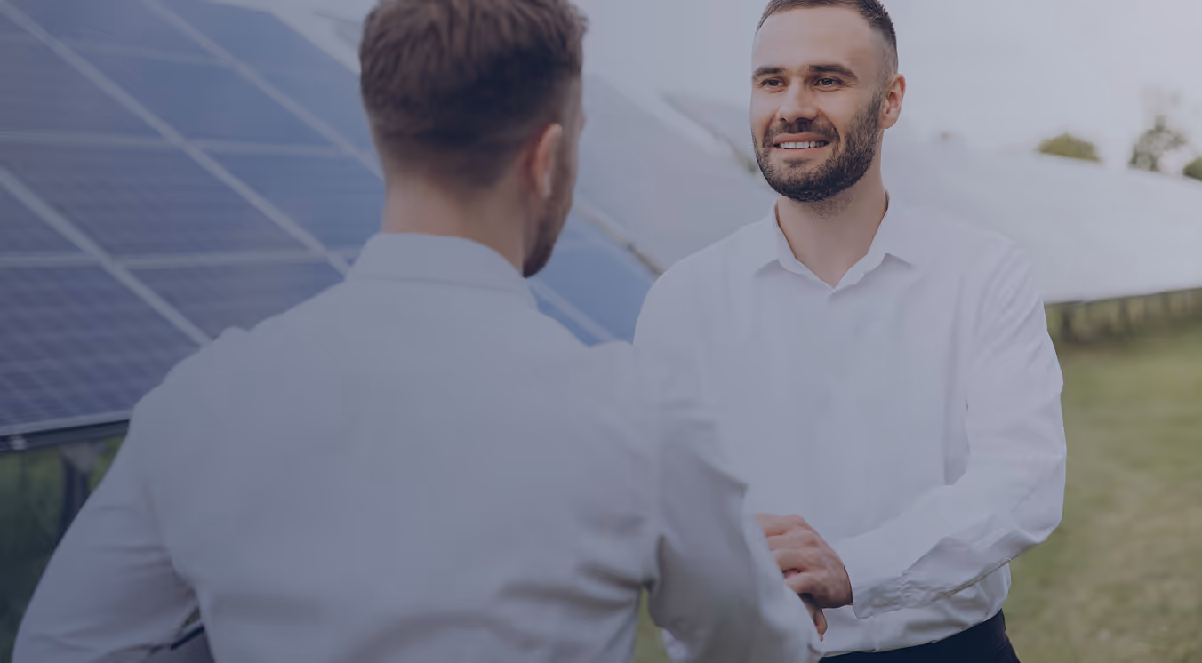 Two men in white shirts shaking hands outdoors near solar panels on a sunny day.