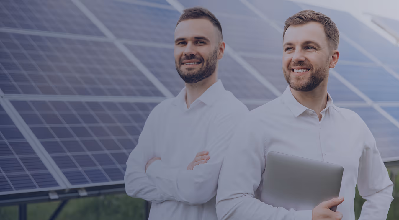 Two men in white shirts smiling and standing in front of solar panels, one holding a laptop.