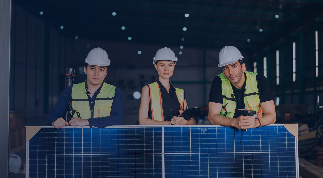 Three engineers wearing safety helmets and reflective vests standing behind a large solar panel in a warehouse.