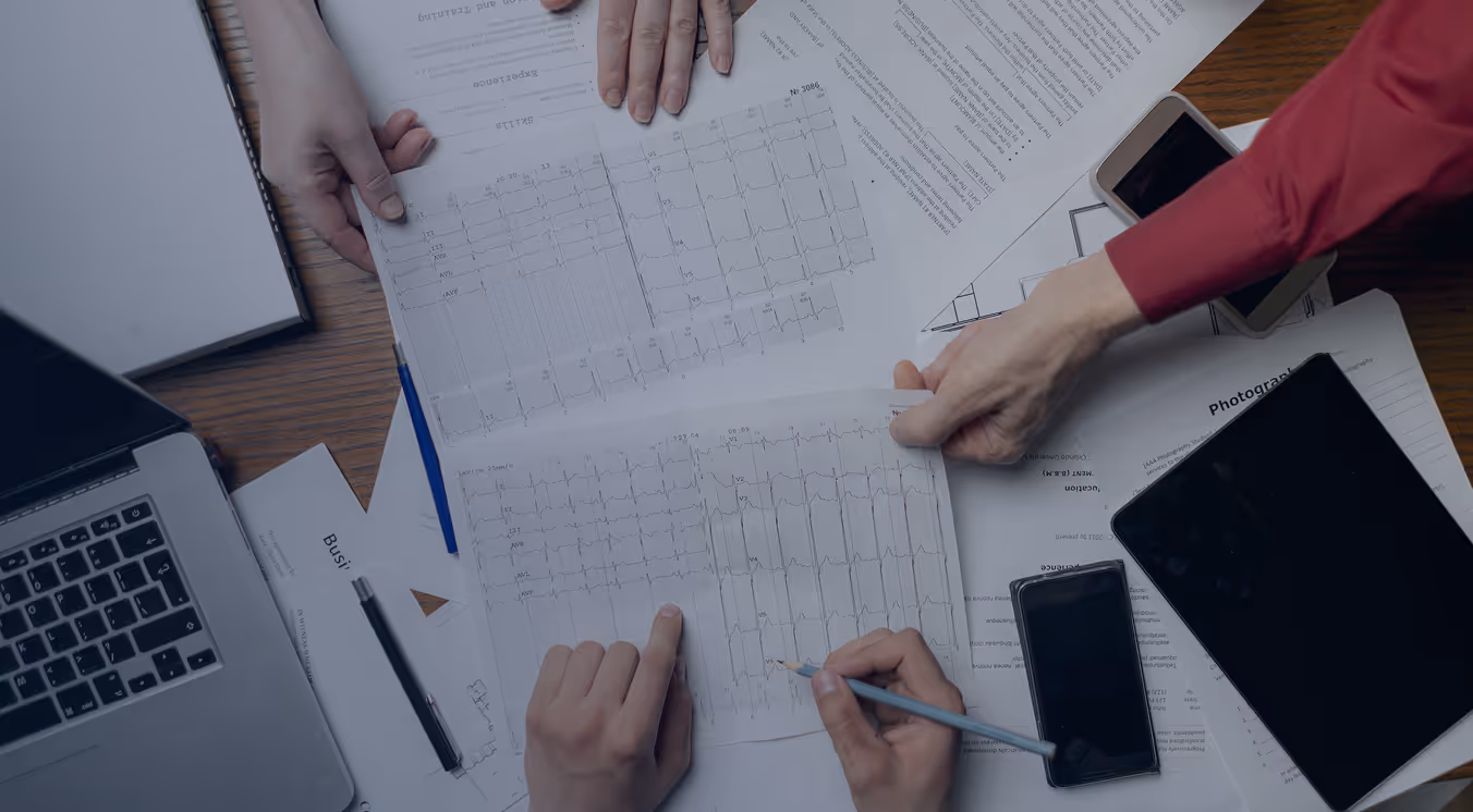 People reviewing and marking electrocardiogram (ECG) charts on a cluttered desk with documents, a laptop, and electronic devices.
