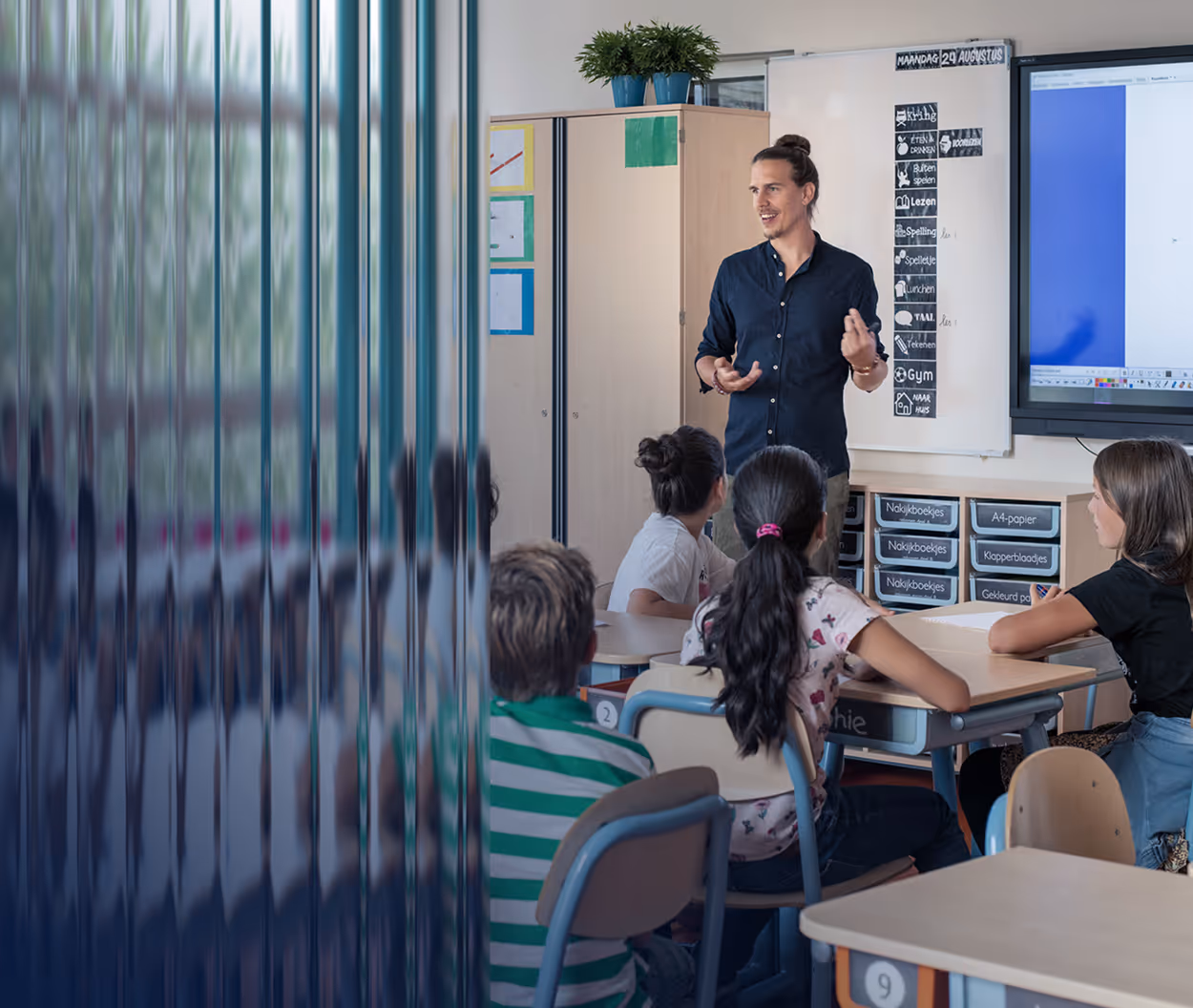 Teacher standing and talking in front of seated students in a classroom with educational posters and a digital screen.