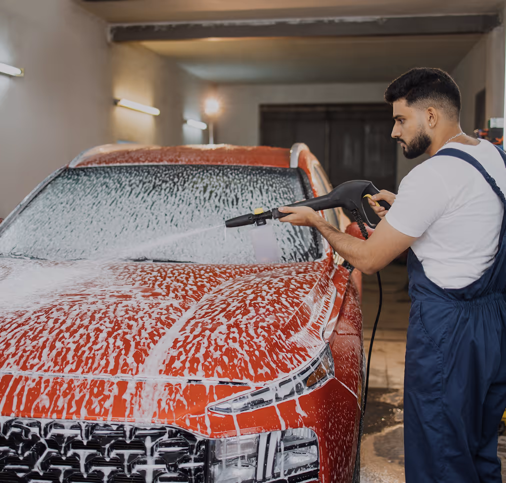 Man in white shirt and blue overalls washing a red car with a high-pressure foam sprayer in an indoor garage.
