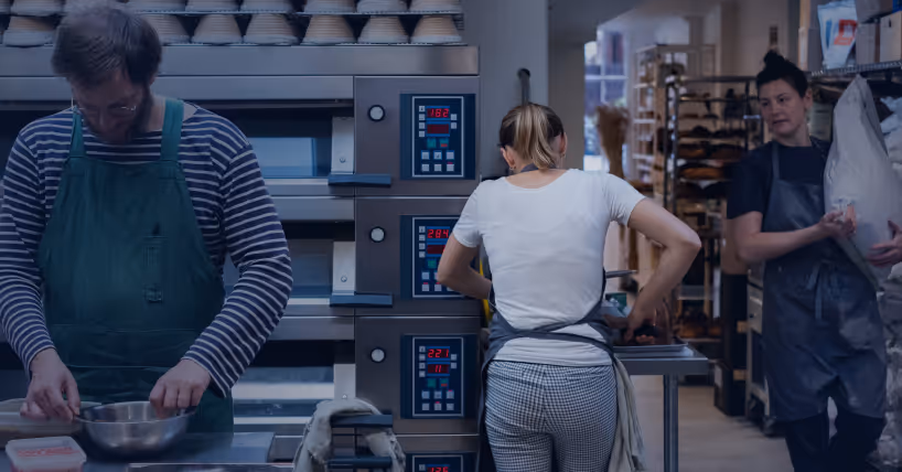 Bakery kitchen with three bakers working; one handling dough in a mixing bowl, one preparing food at a table, and one carrying a large sack of flour.