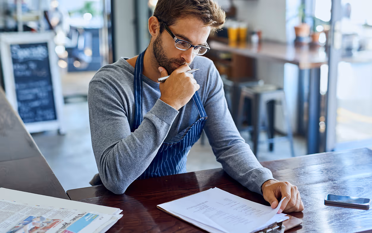 Man wearing glasses and apron thoughtfully reviewing paperwork at a wooden table in a cafe.
