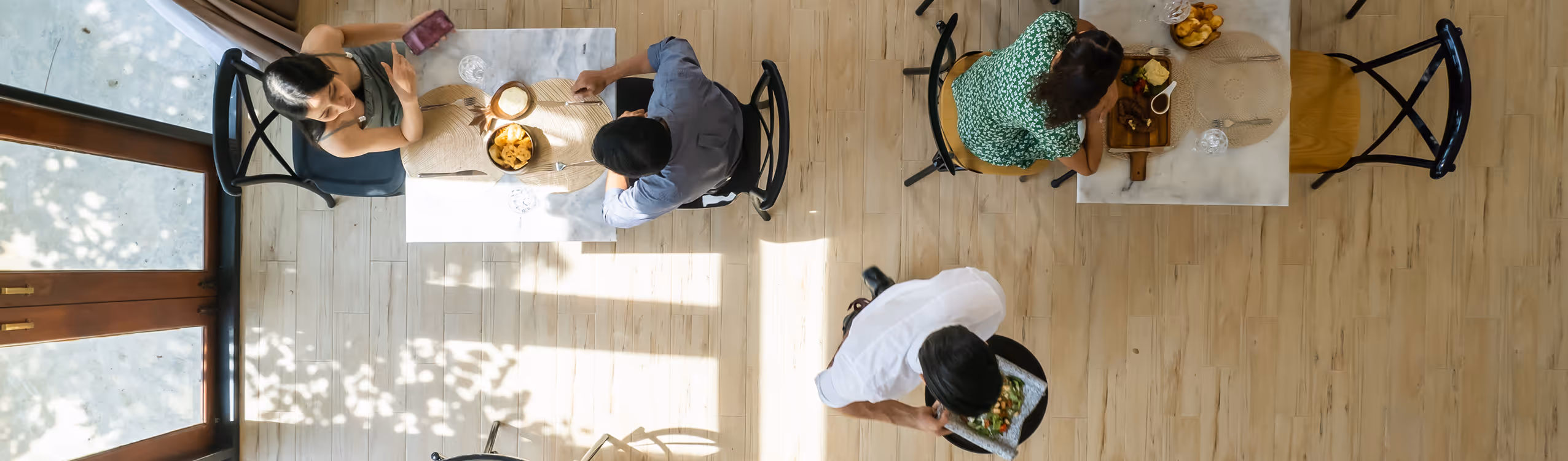Overhead view of two tables with pairs of people seated, a server carrying a plate, and natural light casting shadows on a wooden floor.