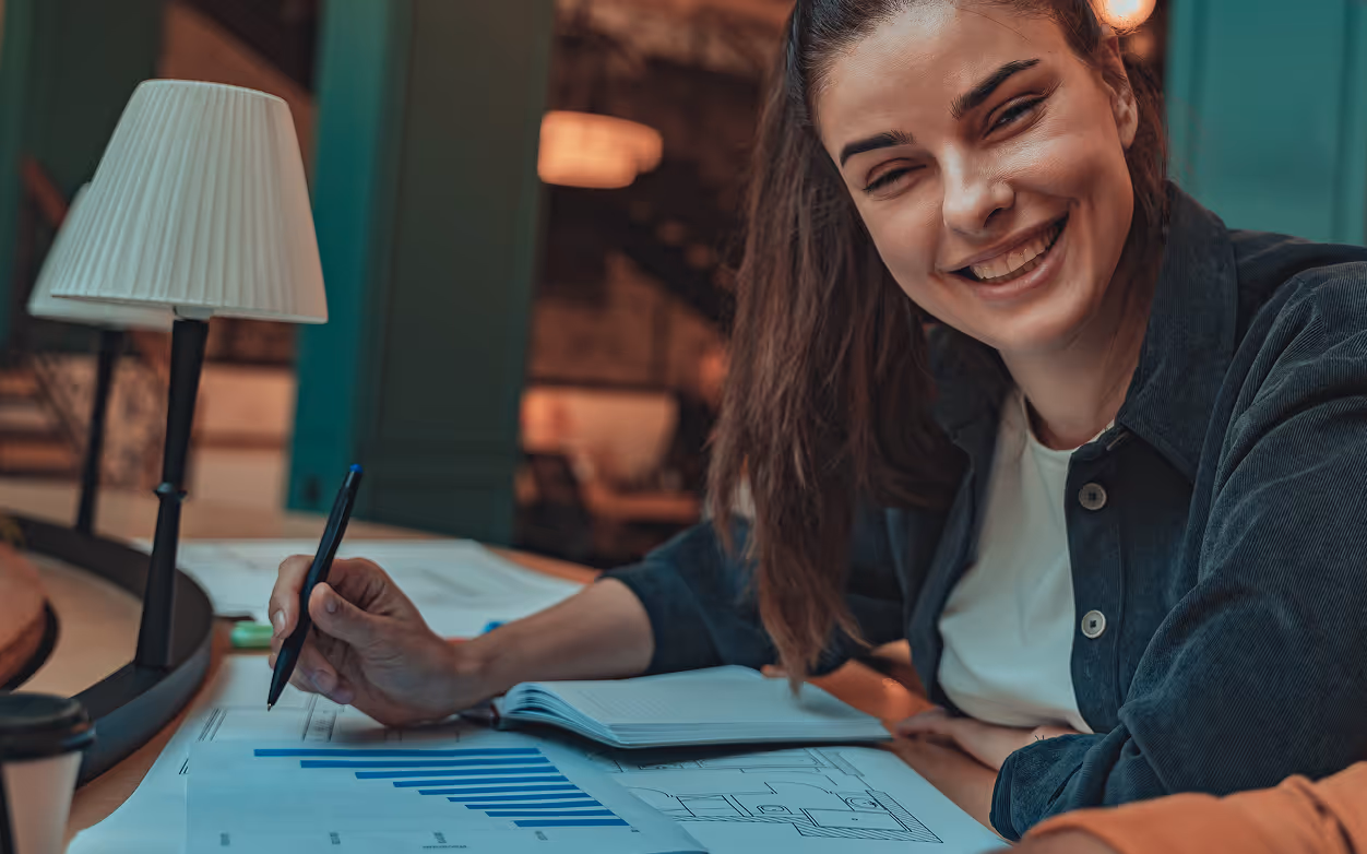 Smiling woman writing on documents with charts at a desk with a lamp and notebook.