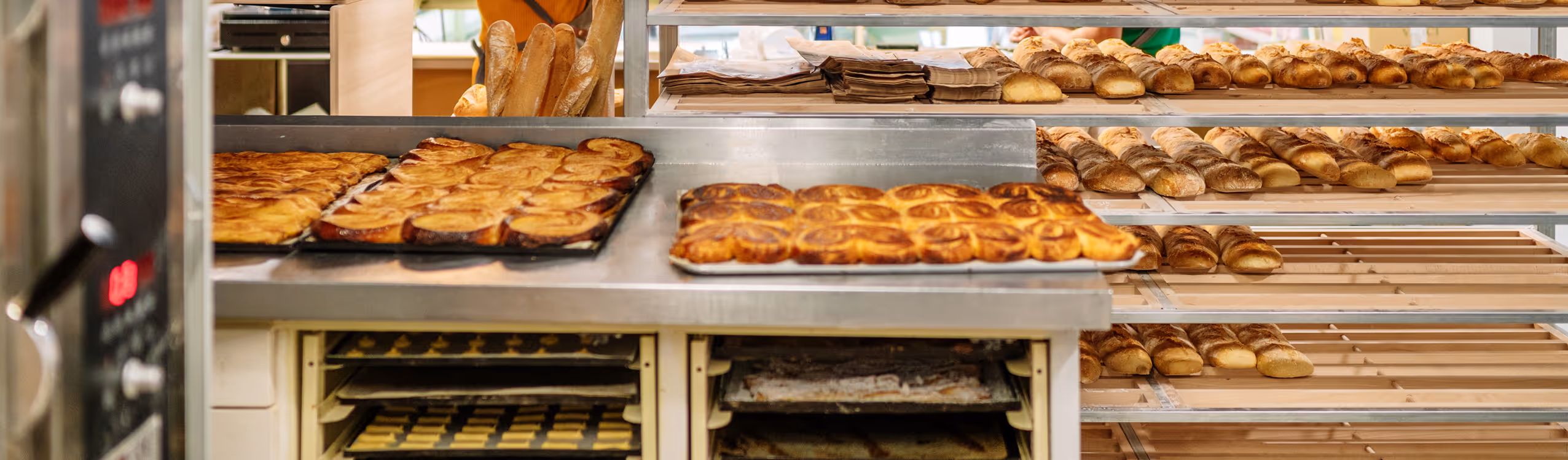 Bakery shelves filled with various types of freshly baked bread and pastries next to an oven with trays of baked goods.