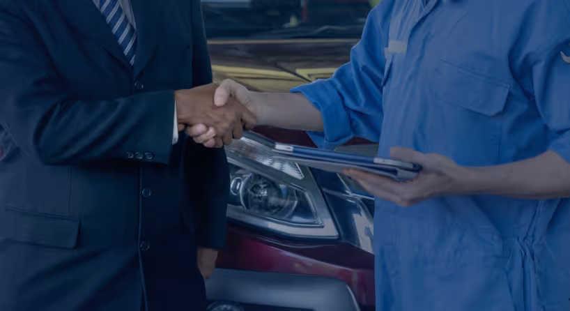 Person in a suit shaking hands with a mechanic holding a clipboard near a vehicle.