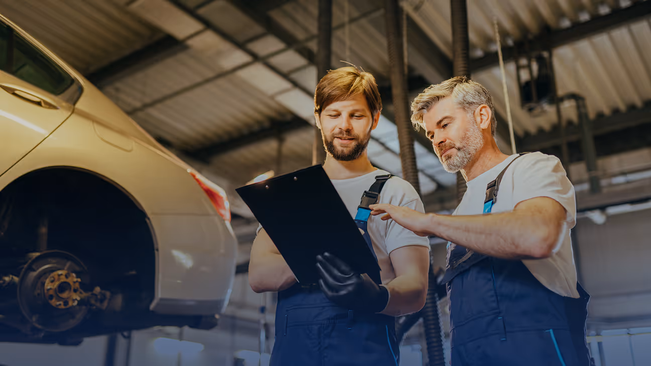 Two mechanics in blue overalls discussing a clipboard next to a car without a rear wheel in a garage.