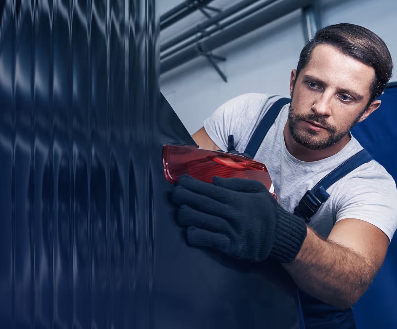 Mechanic wearing black gloves carefully installing a red car tail light on a dark-colored vehicle.
