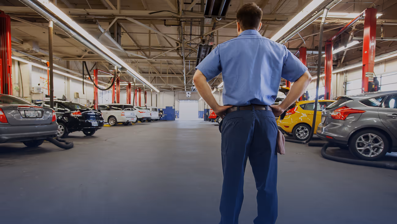 Mechanic in blue uniform standing with hands on hips in a spacious car repair garage with multiple vehicles lined up.