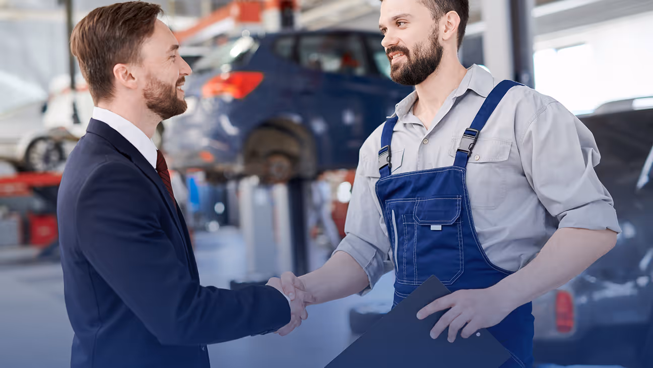 A man in a suit shaking hands with a mechanic in blue overalls inside a car repair shop.