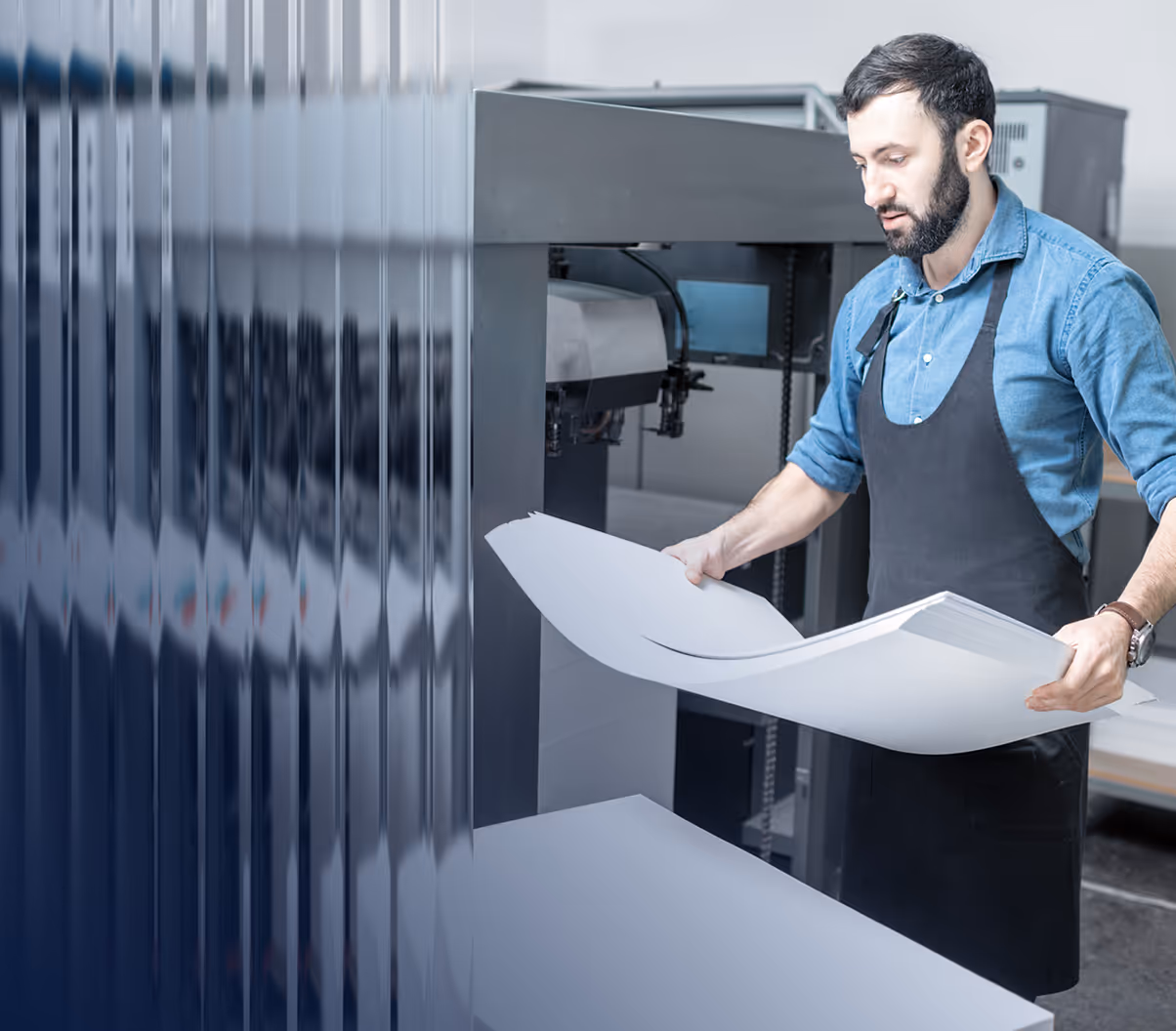 Man in a blue shirt and black apron holding large sheets of paper near a printing machine.