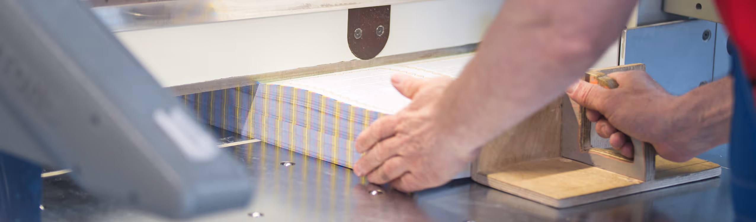 Close-up of a person using a wooden paper cutter to trim a stack of striped paper sheets on a metal table.