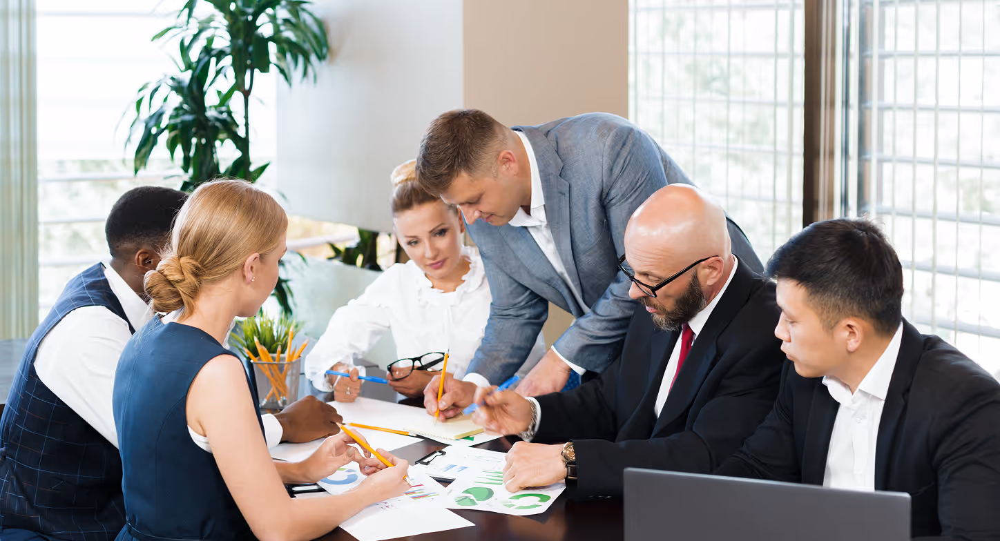 Six business professionals collaborating over documents and charts at a conference table in a modern office.