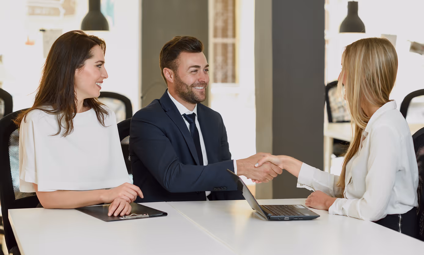 Two businesspeople shaking hands across a table with a third person smiling nearby in a modern office setting.