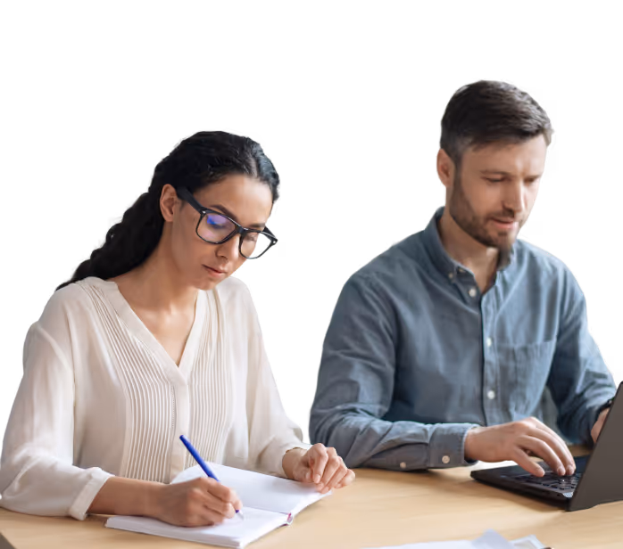 Two colleagues working at a desk, one woman writing in a notebook and one man typing on a laptop.