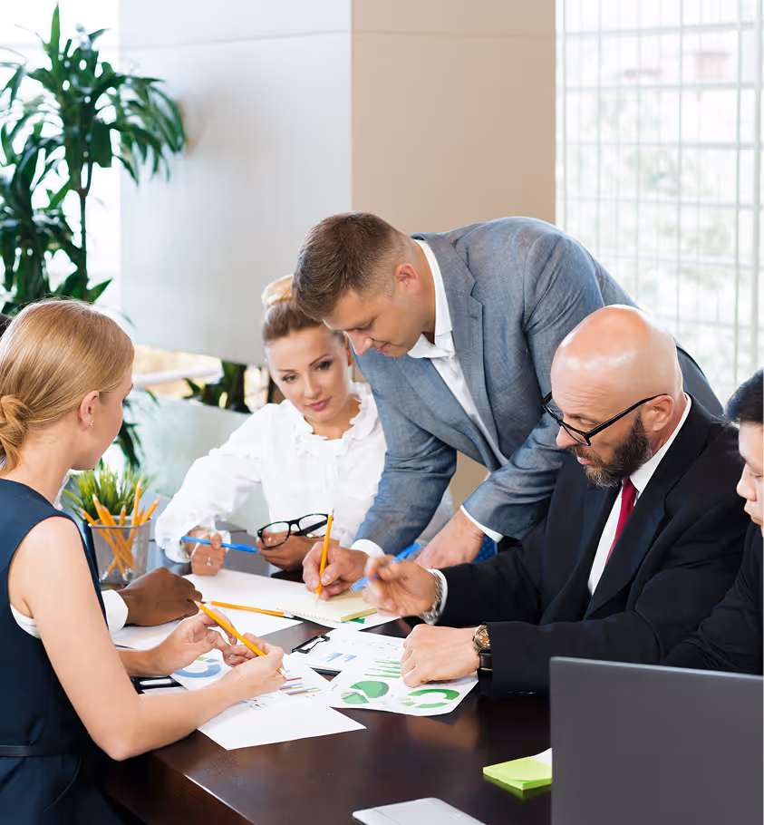 Five business professionals in a meeting discussing charts and taking notes around a table in a modern office.
