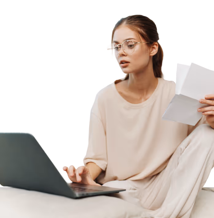 Young woman in glasses sitting with a laptop on her lap, holding papers and typing.