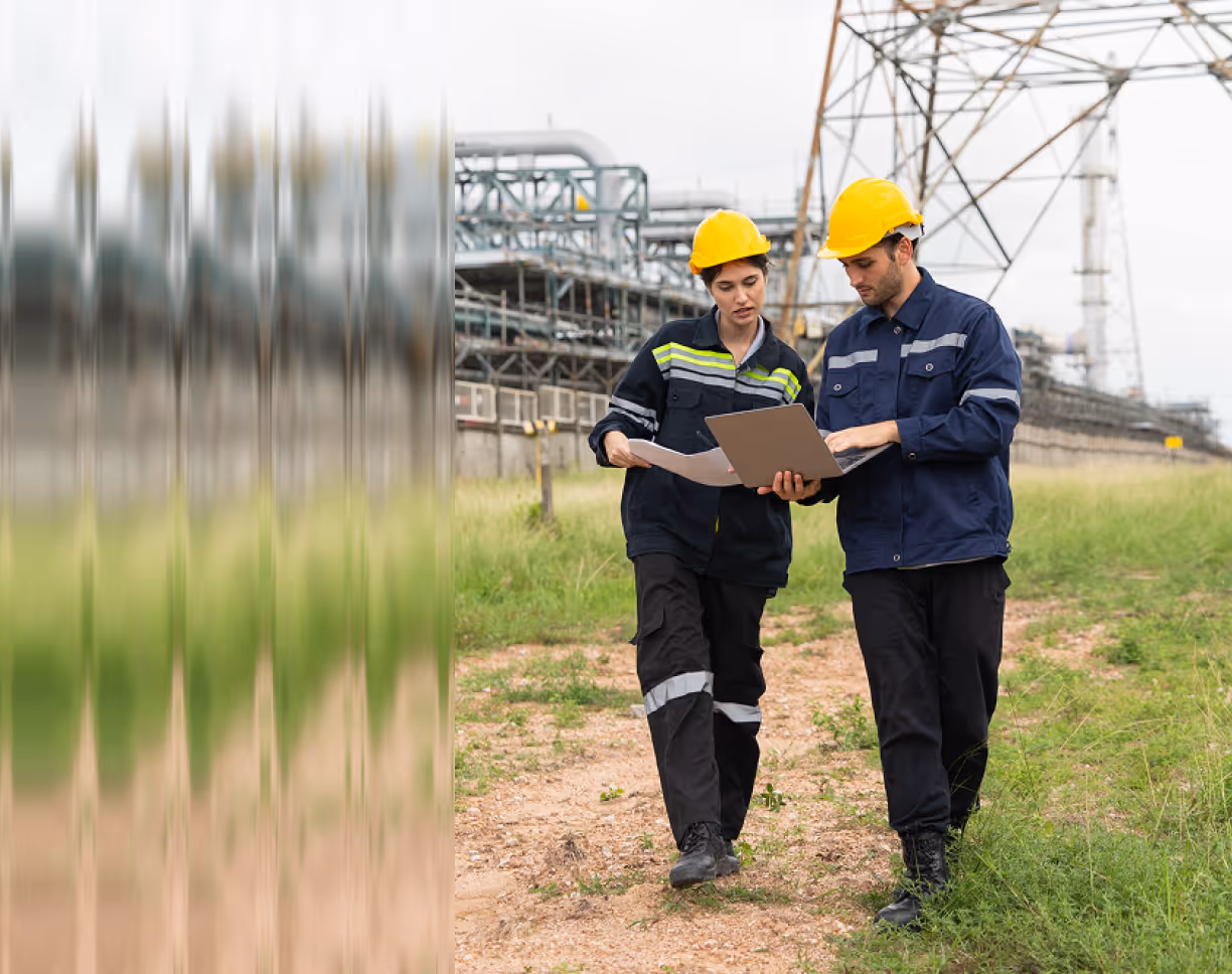 Two engineers in yellow hard hats and navy work uniforms reviewing documents and using a laptop outdoors at an industrial site.