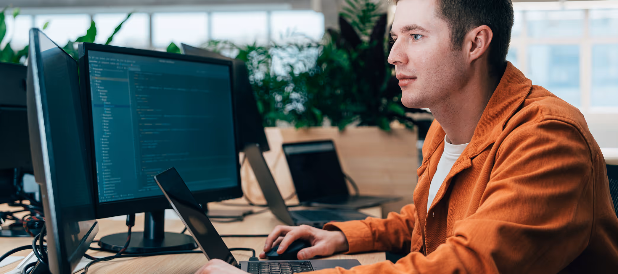 Man in orange jacket working on laptop with dual monitors displaying code in a bright office.