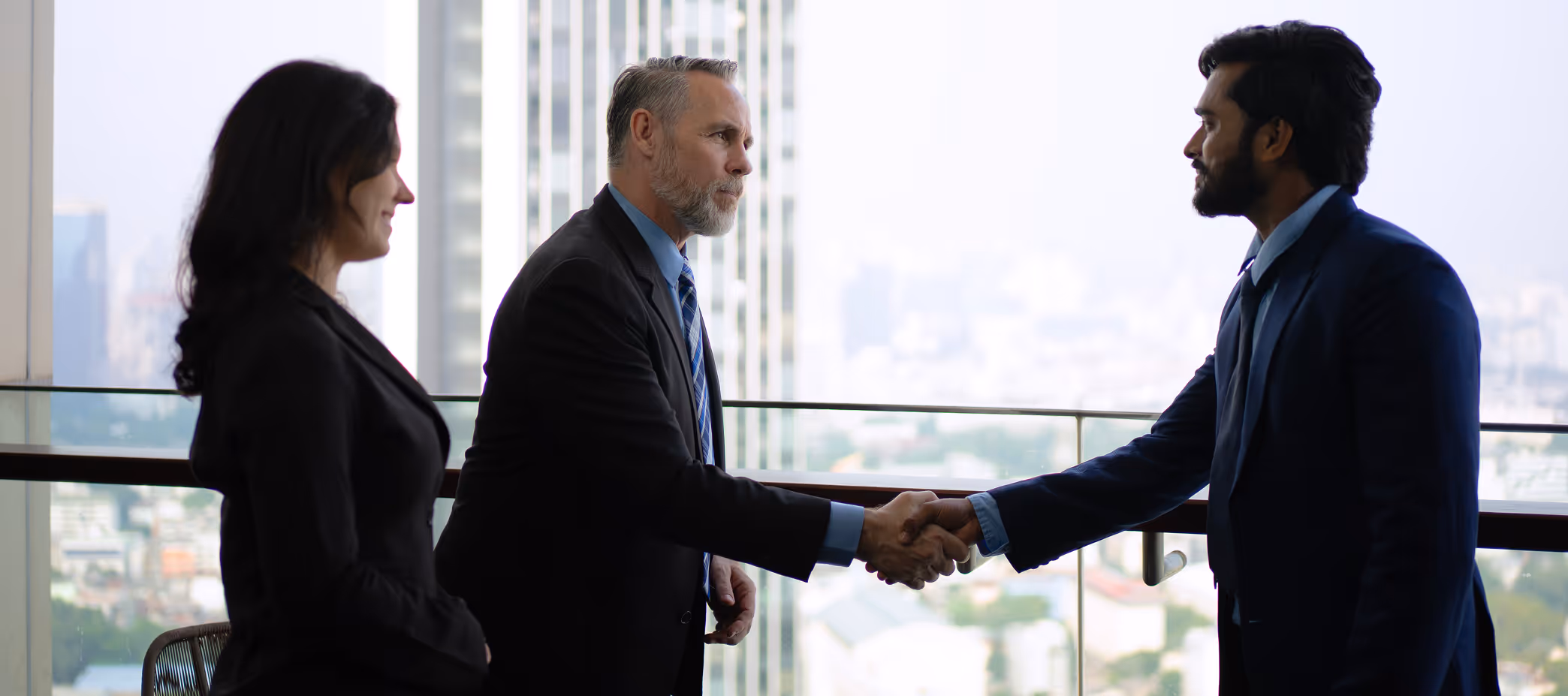 Two men in business suits shaking hands while a woman in business attire stands nearby with a cityscape in the background.