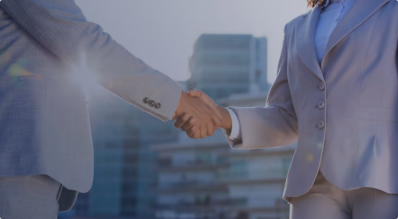 Two business professionals in suits shaking hands outdoors with modern buildings in the background.