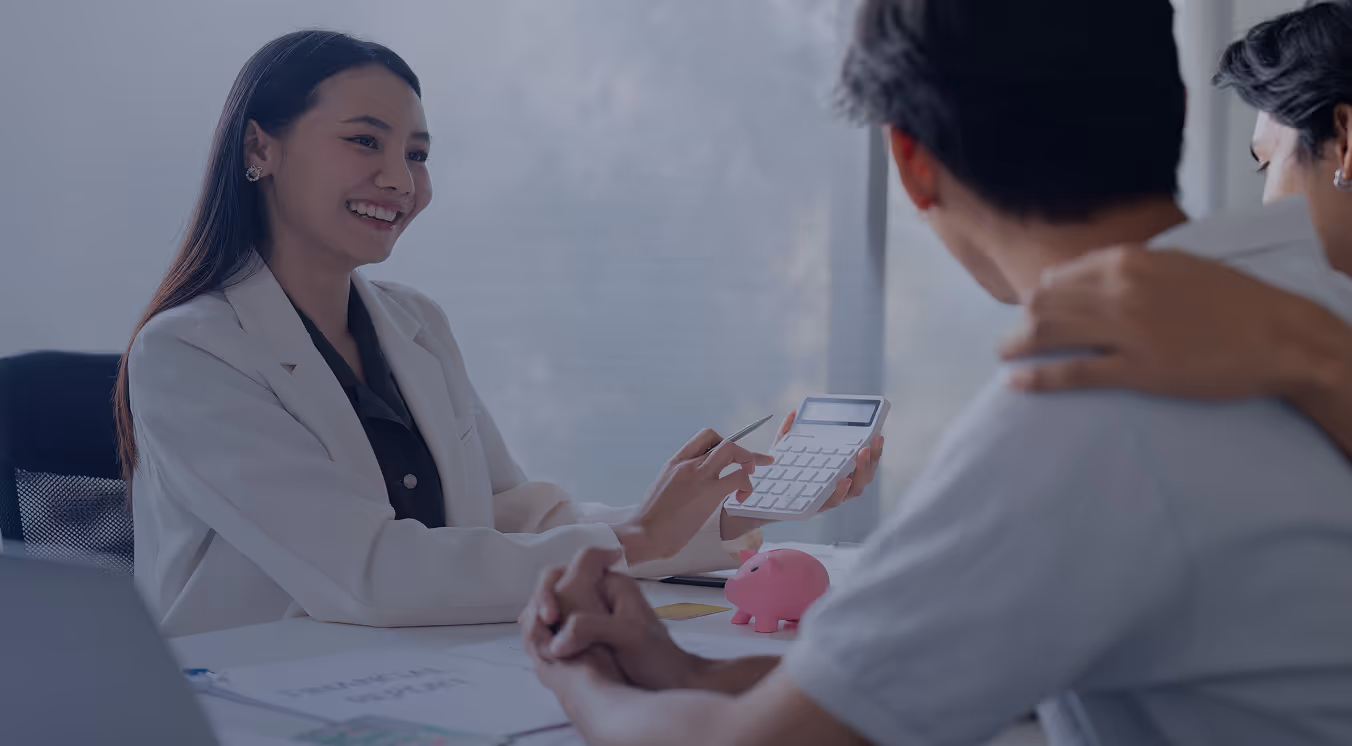 Smiling female financial advisor showing a calculator to a couple discussing finances at a desk with a pink piggy bank.