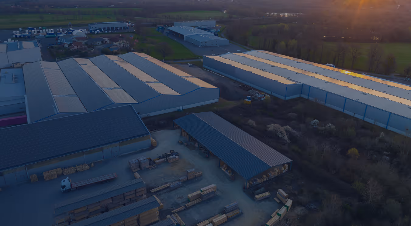 Aerial view of industrial warehouses and storage area with stacks of wooden pallets and materials at sunset.