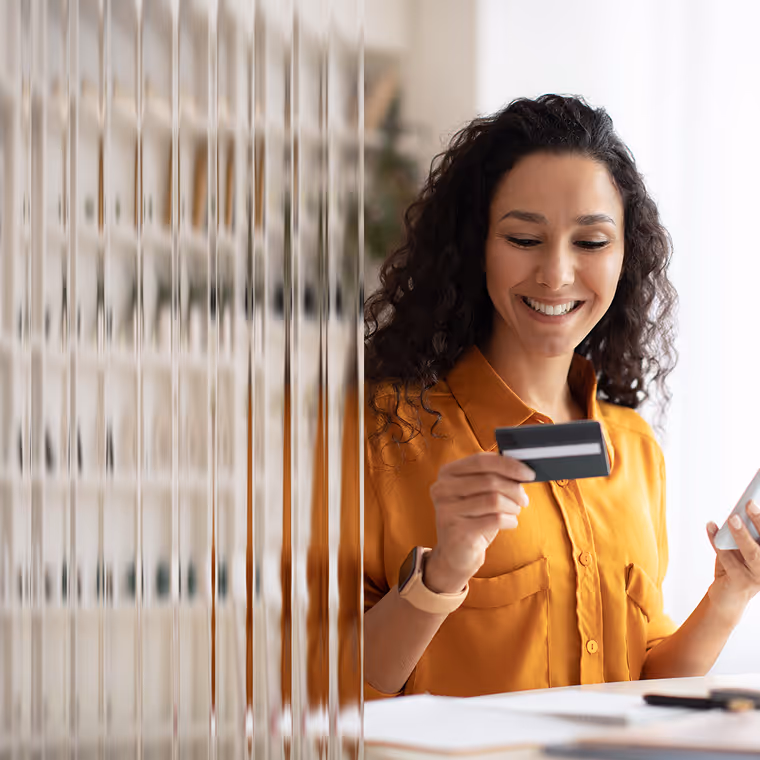 Smiling woman holding a credit card in one hand and a smartphone in the other, standing indoors near a blurred glass panel.
