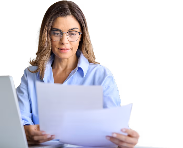 Woman wearing glasses and a blue shirt sitting at a desk, reviewing documents with a laptop nearby.