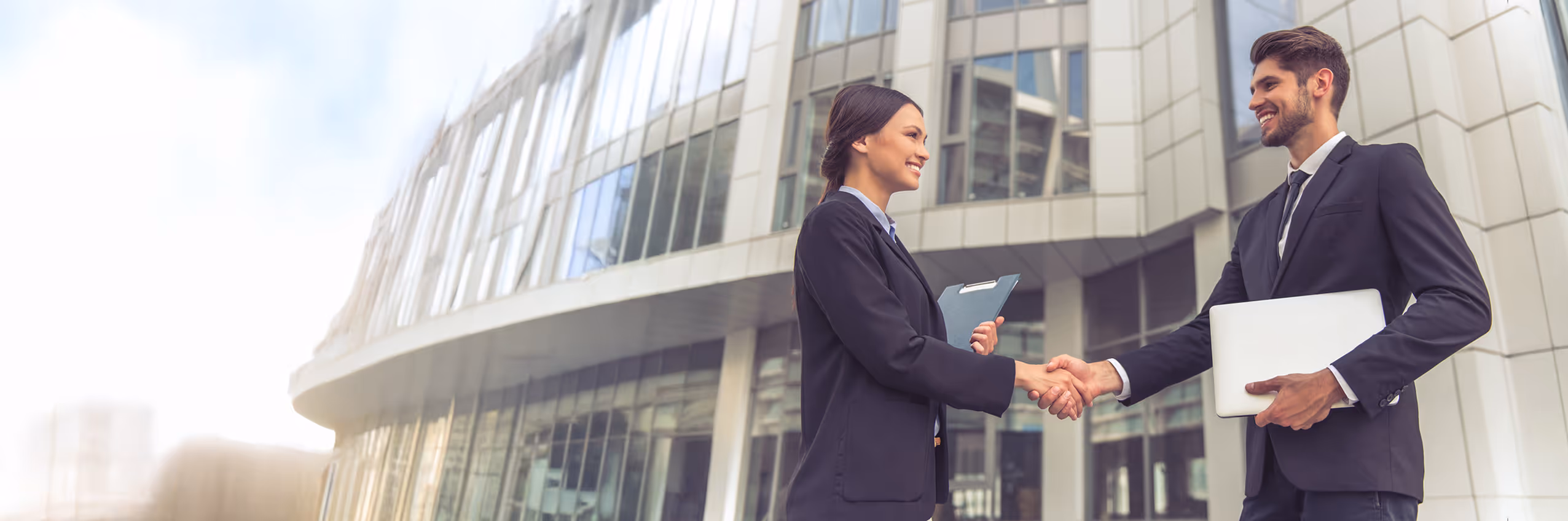 Two business professionals shaking hands outside a modern office building, one holding a laptop and the other a clipboard.