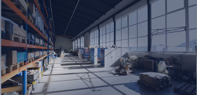 Bright spacious industrial warehouse with tall shelves and large windows casting shadows on the floor.