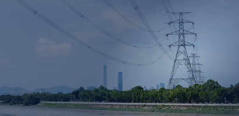 Power transmission towers with cables over a green riverside park and a city skyline in the background under a clear sky.