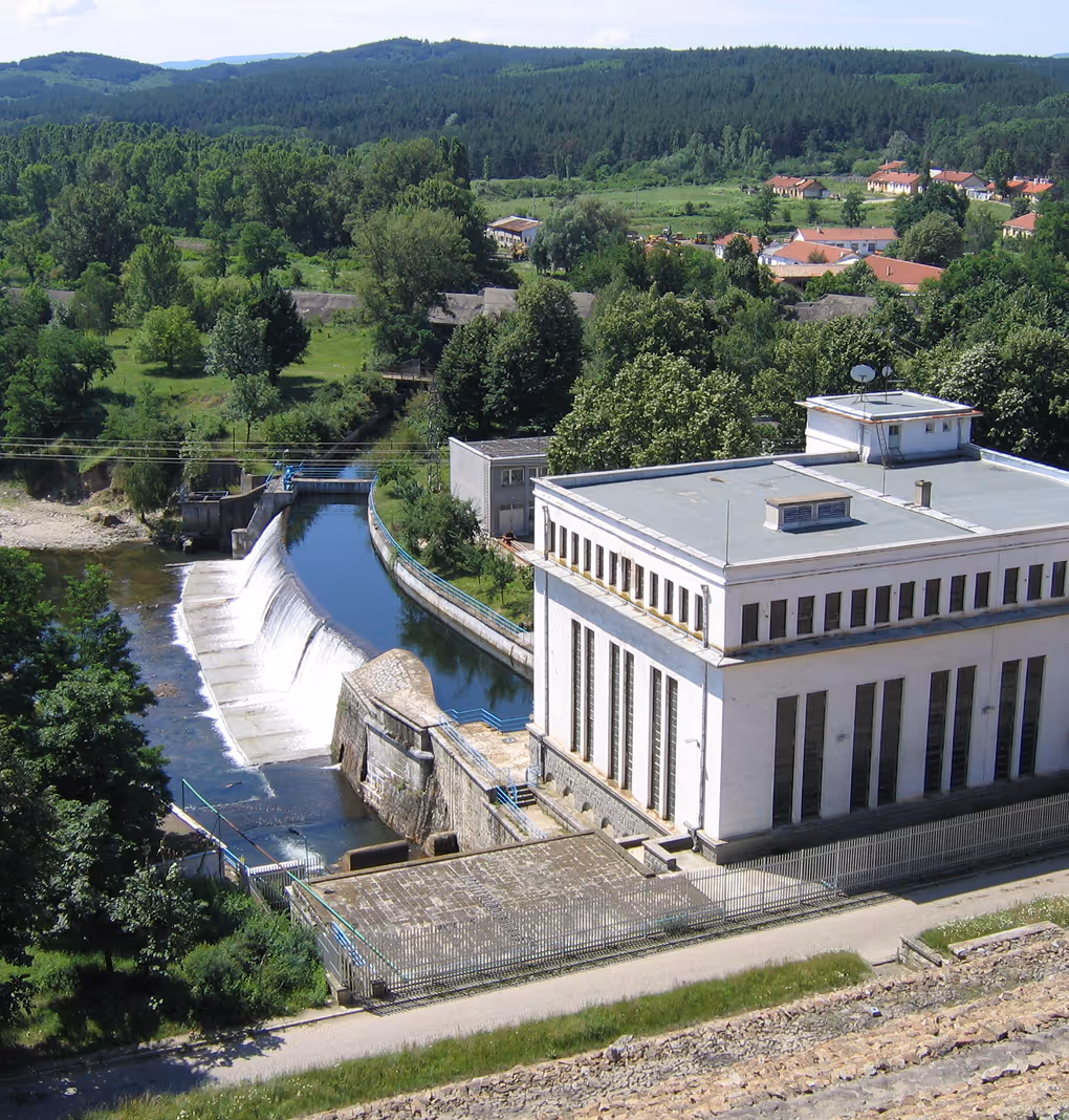 A small hydroelectric dam with water flowing over the spillway, surrounded by dense green trees and hills in the background.