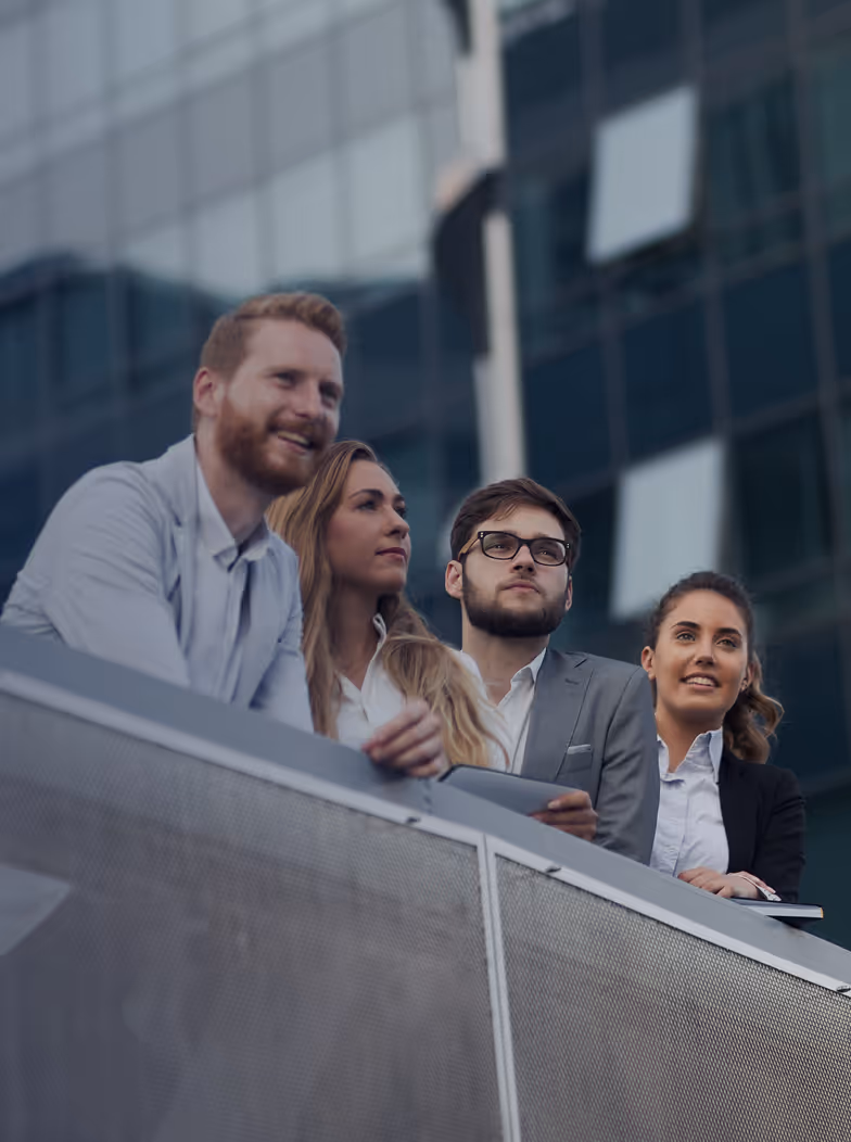 Group of four business professionals standing outdoors in front of a modern glass building, looking thoughtfully into the distance.