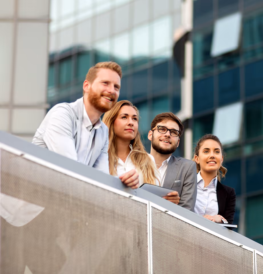 Four young professionals standing outdoors, leaning on a railing with a glass office building in the background, looking forward thoughtfully.